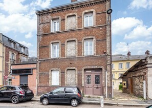 Un edificio de varios pisos de ladrillo con una puerta de garaje en la planta baja, un coche negro estacionado frente a él, y edificios más antiguos aledaños.
