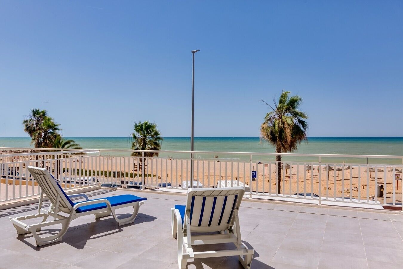 A terrace with two sun loungers, a clear view of the beach, and a palm tree.
