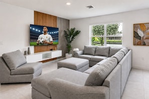 Ein modernes Wohnzimmer mit einem Ecksofa, einem Flachbildfernseher, einer Wandpaneele aus Holz und einem großen Fenster mit Blick auf Grünflächen.