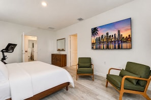Ein Hotelzimmer mit einem Bett, zwei Stühlen, einem Schreibtisch mit Computer, einem Spiegel und einem Stadtbild an der Wand.