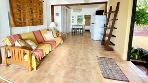 Ein Wohnzimmer mit einer Couch, einem Couchtisch, einem Esstisch und einer Küchenzeile inklusive Kühlschrank und Regalen.