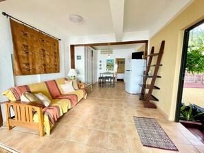 Ein Wohnzimmer mit einer Couch, einem Couchtisch, einem Esstisch und einer Küchenzeile inklusive Kühlschrank und Regalen.
