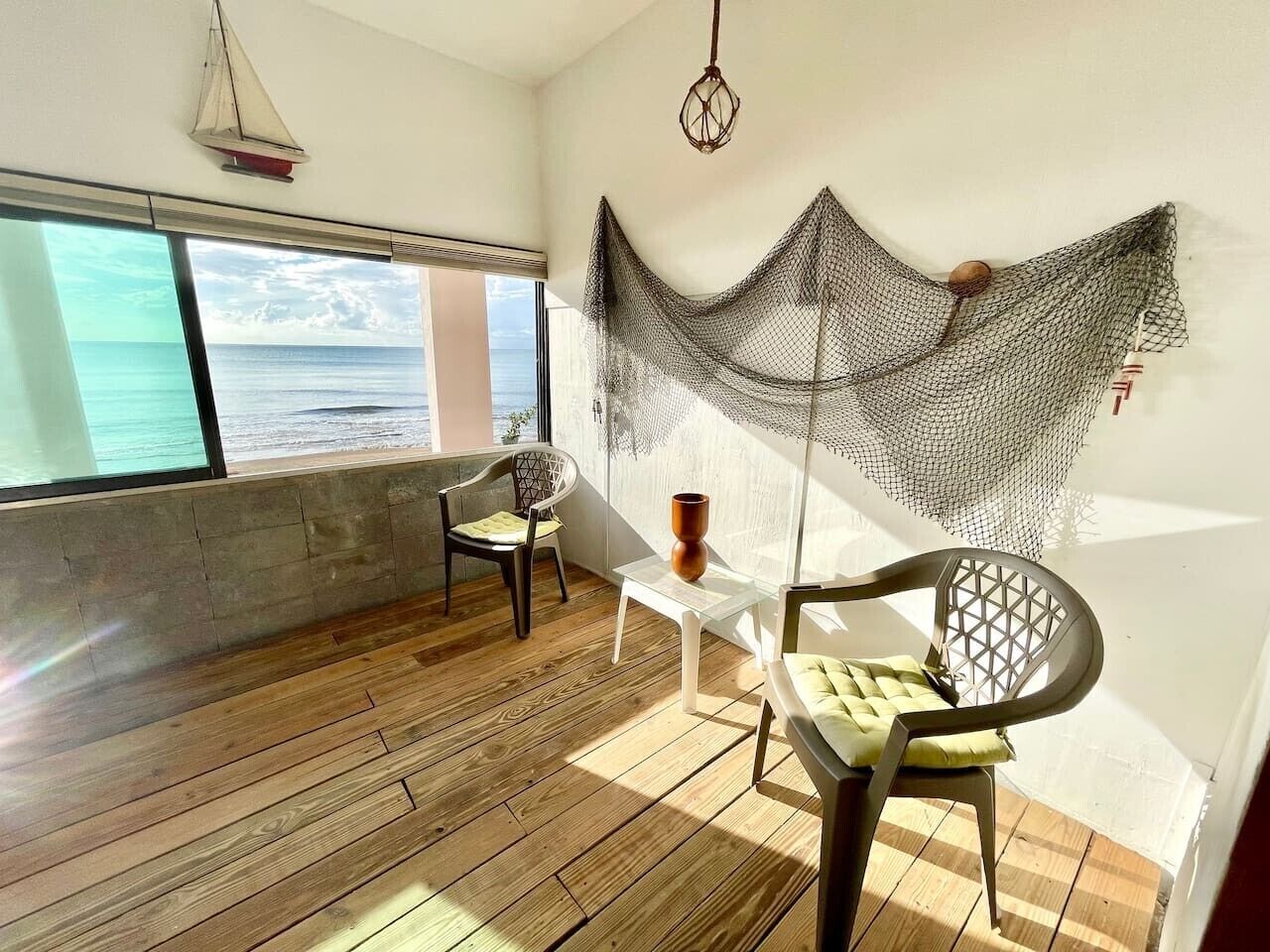Una habitación con piso de madera, dos sillas, una mesita y una red decorativa, con vista al mar a través de una ventana.