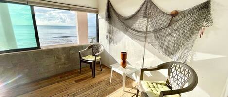 Una habitación con piso de madera, dos sillas, una mesita y una red decorativa, con vista al mar a través de una ventana.