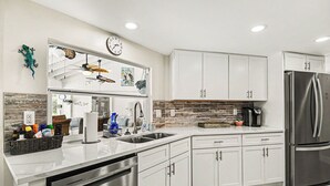 A modern kitchen with white cabinets, stainless steel appliances, a clock, and decorative wall art. - Palaco Paradise - Waterfront Oasis with Large Pool (Cape Coral)