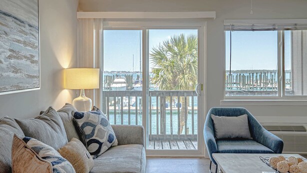 Un salón con sofá, sillón y una mesa con adornos. Tiene un balcón con vistas al agua y palmeras.