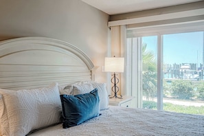 A bed with a headboard, pillows, a bedside table with a lamp, and a view of a pool and greenery through a window. - Waterfront Condo with Marina Views in Panama City (Panama City)