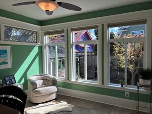 Ein Zimmer mit grüner Wand, Deckenventilator, Fenster mit Blick auf ein Haus, ein weißer Sessel und ein kleiner Tisch mit einer Pflanze.