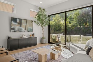 Ein modernes Wohnzimmer mit großem Fernseher, einem grauen Schrank, einer Zimmerpflanze, einer Couch und einem großen Fenster mit Blick auf Bäume.
