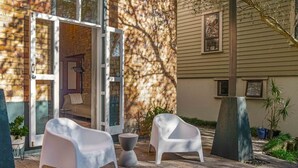 Un patio con dos sillas blancas, una mesita y un ventanal de vidrio que da a un espacio interior con un sofá.