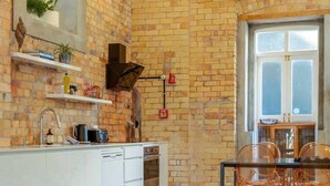 Una cocina moderna con pared de ladrillo, muebles blancos y un comedor con mesa y sillas de vidrio.