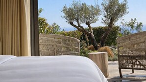 A hotel room with a bed, wicker chairs, and a view of olive trees and greenery.