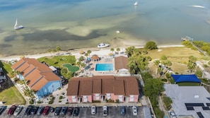 An aerial view of a coastal hotel with a swimming pool, beach access, and parked cars. - Cozy St. Pete Condo |  Pool | Near Beaches (St. Petersburg)
