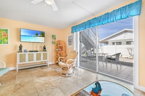 A living room with a large TV, a wicker chair, a glass table, and a view of a house through a sliding glass door. - Cozy St. Pete Condo |  Pool | Near Beaches (St. Petersburg)