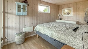 Makuuhuone, jonka seinät ovat puuta, siinä on ikkuna, kuviollinen päiväpeite päällystetty sänky, kaappi ja patteri.