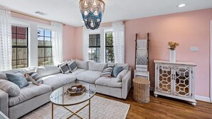 A living room with a sectional sofa, a glass coffee table, a white cabinet with a geometric design, and a decorative chandelier. - Blue Haven | Hot Tub, Fire Pit & Outdoor Oasis (Galveston)