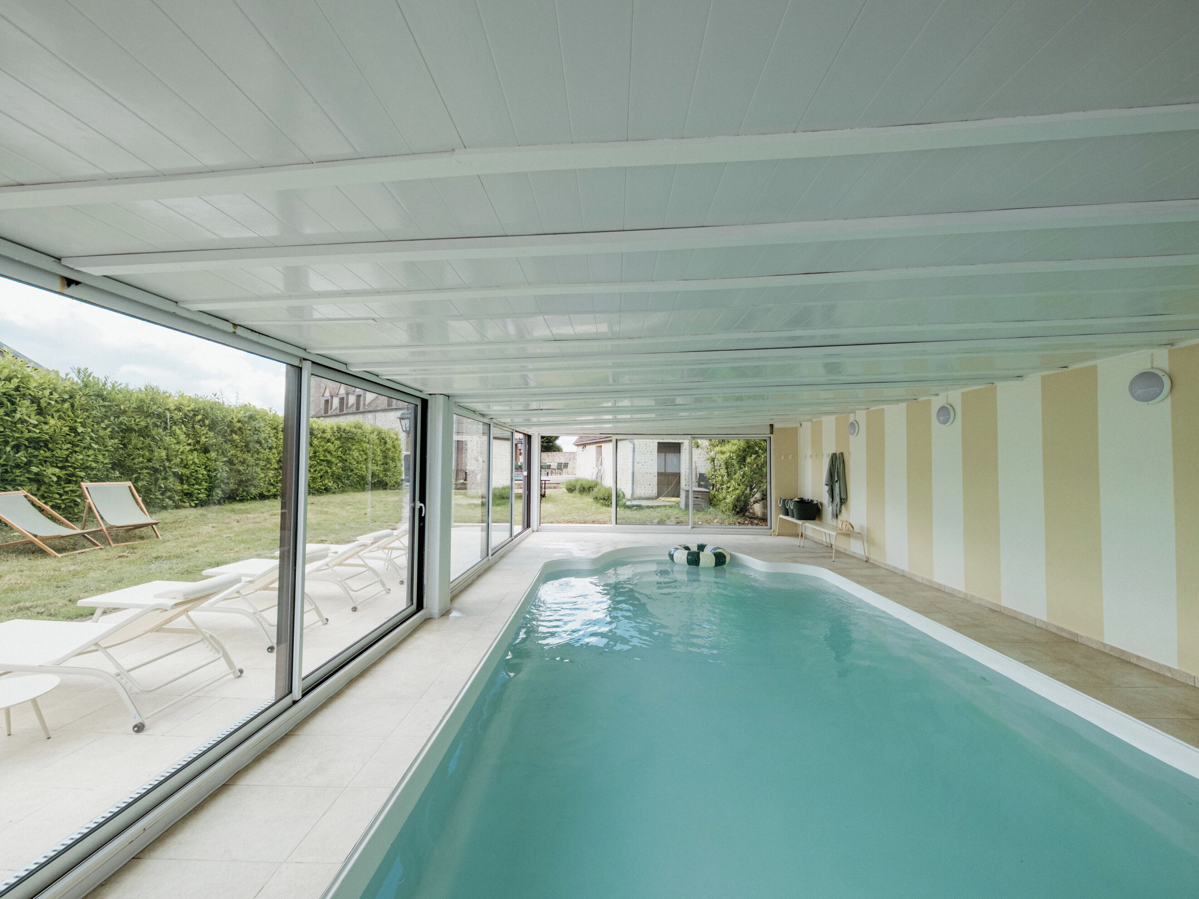 An indoor swimming pool with clear glass walls, lounge chairs, and a view of the outside.