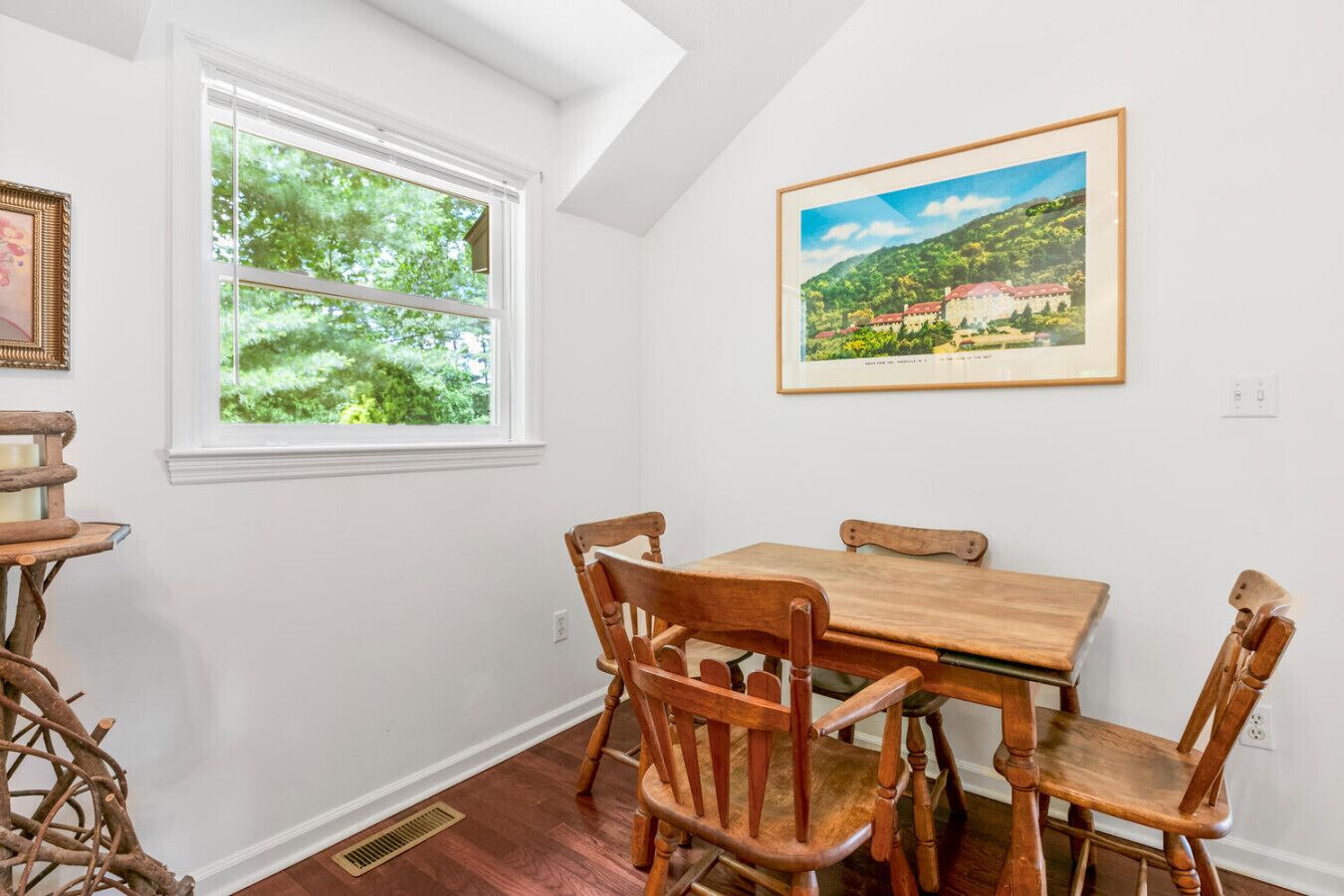 Ein Essbereich mit Holztisch und Stühlen, einem Fenster mit Blick ins Grüne und einem gerahmten Bild an der Wand.