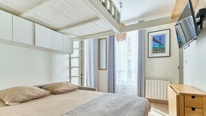 A bedroom with a bed, a wooden dresser, a television, a radiator, and a window with curtains. - Opera · 9th · Cosy Studio with Mezzanine – managed by Mey Agency (Paris)