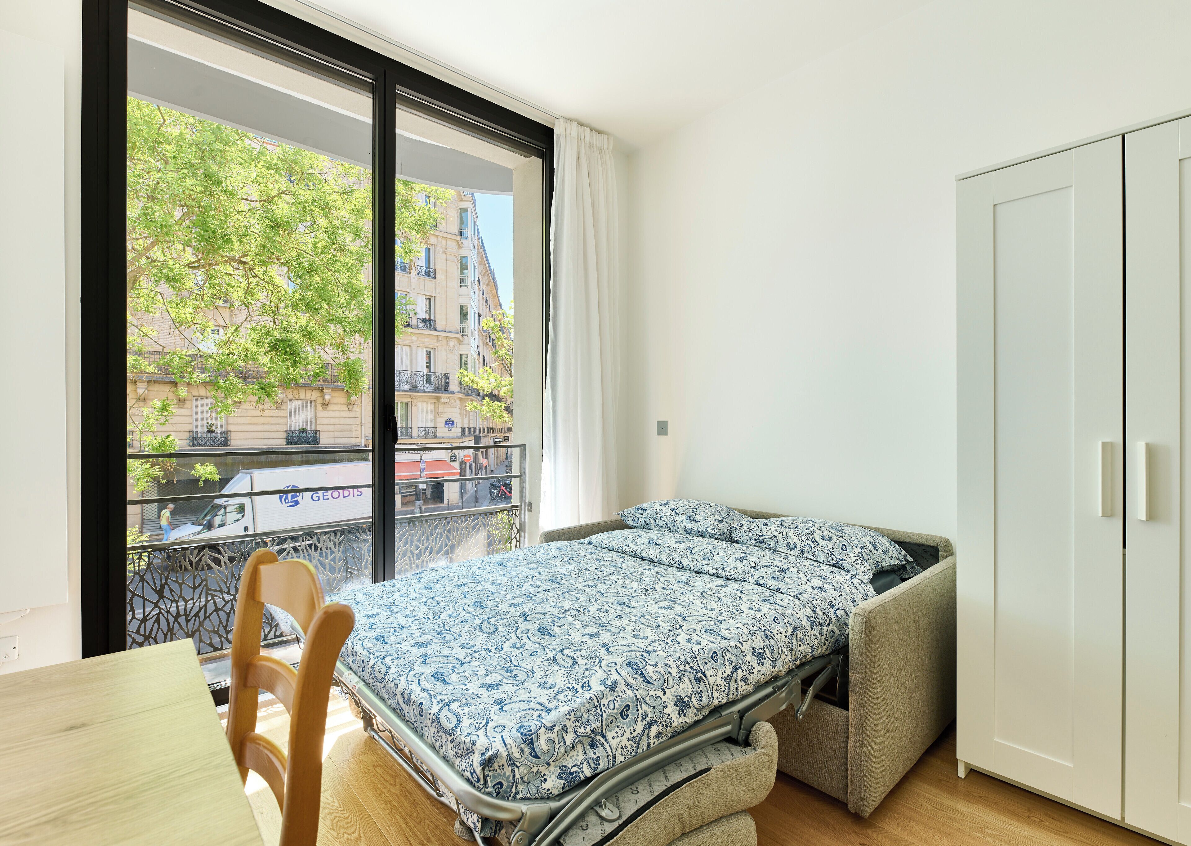 A bedroom with a bed, a chair, a wardrobe, and a window with a view of buildings and trees.