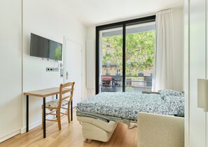 A modern bedroom with a bed, a desk, and a chair, a television, and a large window with a view of buildings and trees. - Auteuil · 16th · Bright & Minimalist Studio – managed by Mey Agency (Paris)