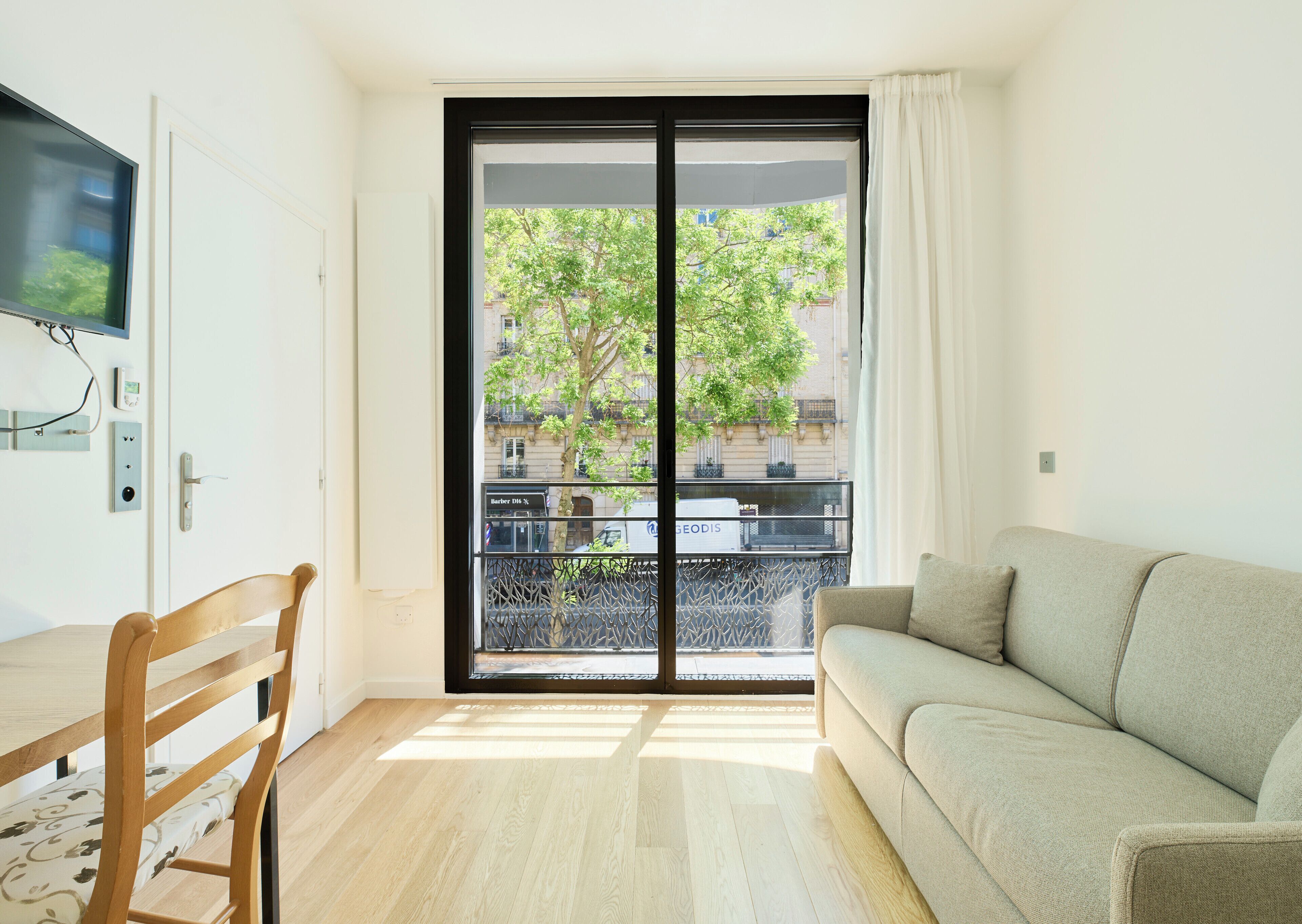 A room with a sofa, a wooden chair, a TV, a sliding glass door, and a view of buildings and trees.