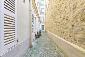 Une ruelle étroite au sol pavé, avec des volets blancs et un mur à la texture particulière.