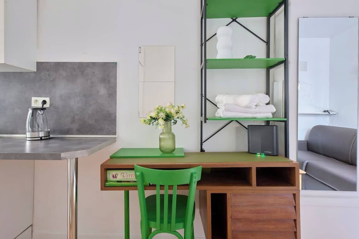 Una habitación con escritorio, silla, estanterías verdes, un jarrón con flores, máquina de café y sofá gris.