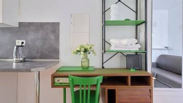 Una habitación con escritorio, silla, estanterías verdes, un jarrón con flores, máquina de café y sofá gris.