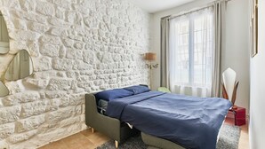 Un dormitorio con pared de piedra, una cama con ropa de cama azul, una lámpara de noche y una ventana con cortinas.