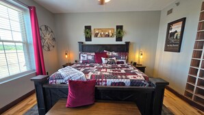 Une chambre à coucher comprenant un lit, des tables de chevet, une commode, un ventilateur de plafond, une fenêtre avec des stores et un mur orné d’une horloge et d’un tableau encadré.