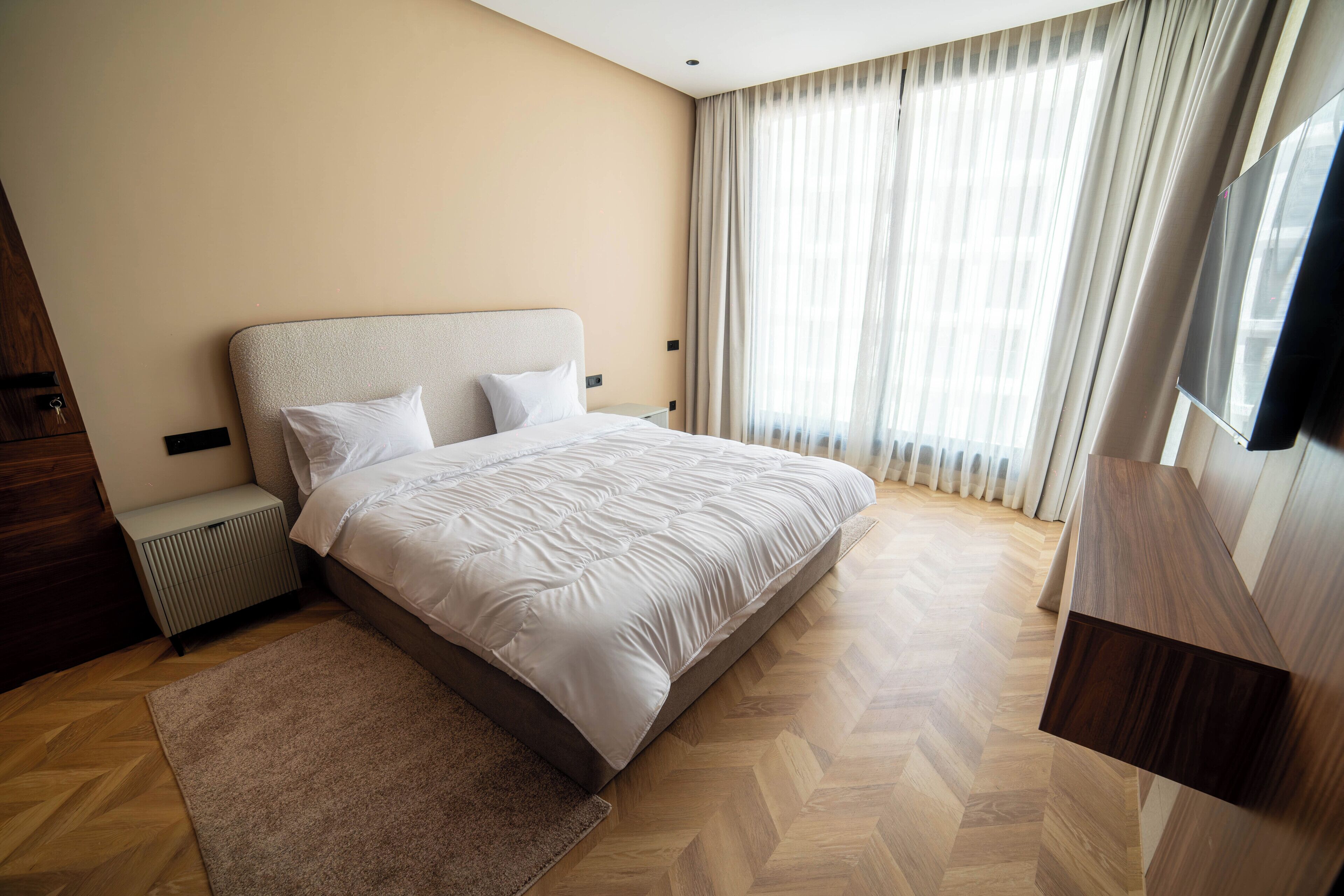 A modern hotel room with a large bed, a wooden floor, a TV, and a window with curtains.