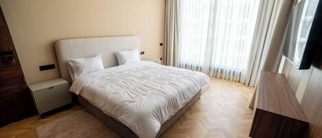 A modern hotel room with a large bed, a wooden floor, a TV, and a window with curtains.
