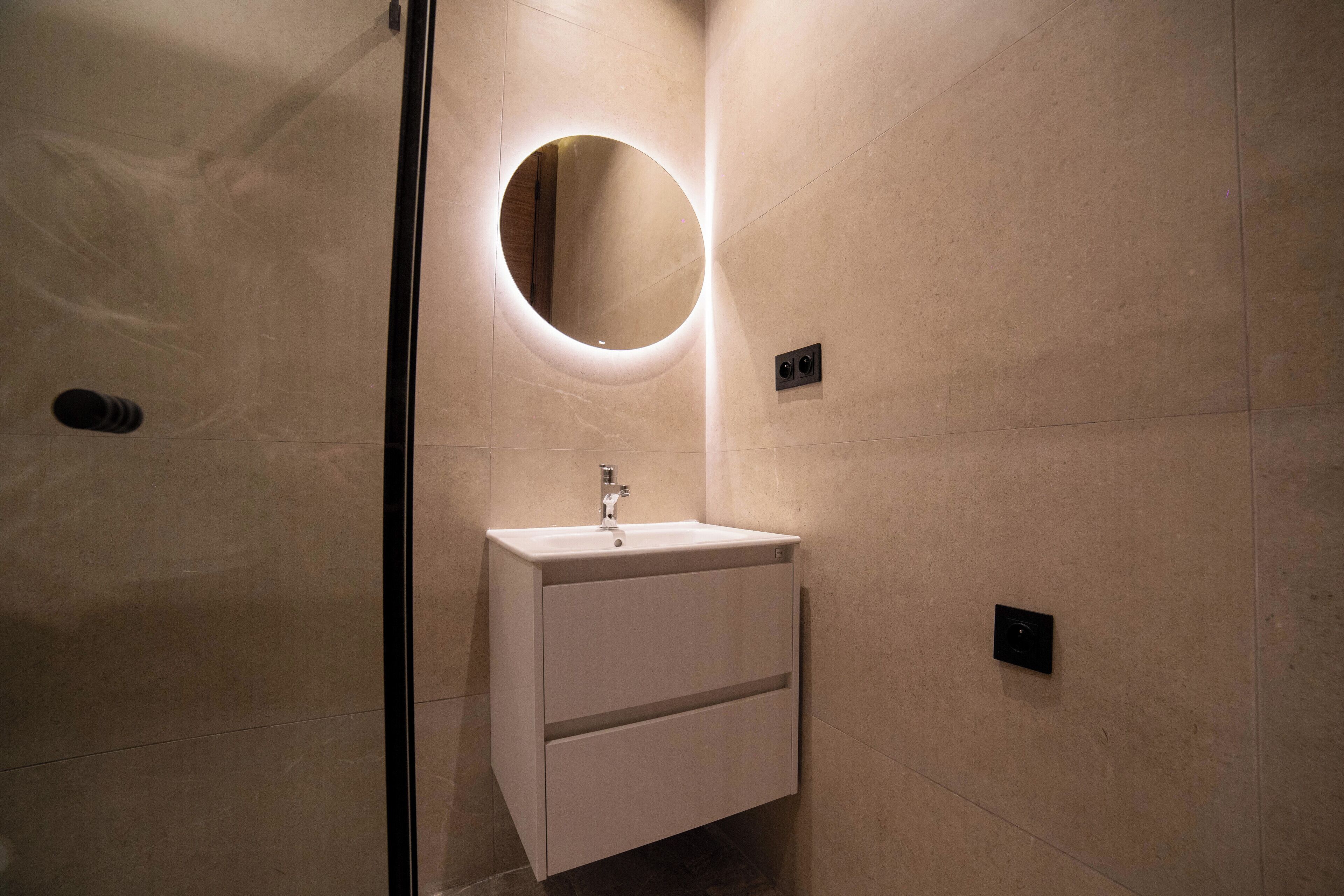 Une salle de bain moderne avec un miroir rond, un lavabo blanc et un luminaire fixé au mur.