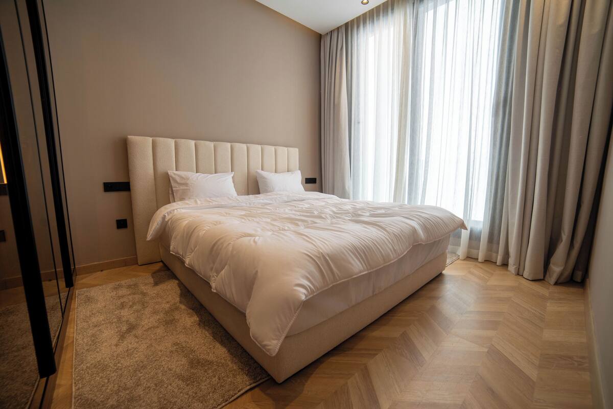 Un dormitorio moderno con una cama grande, cabecero y vistas al exterior a través de cortinas traslúcidas.