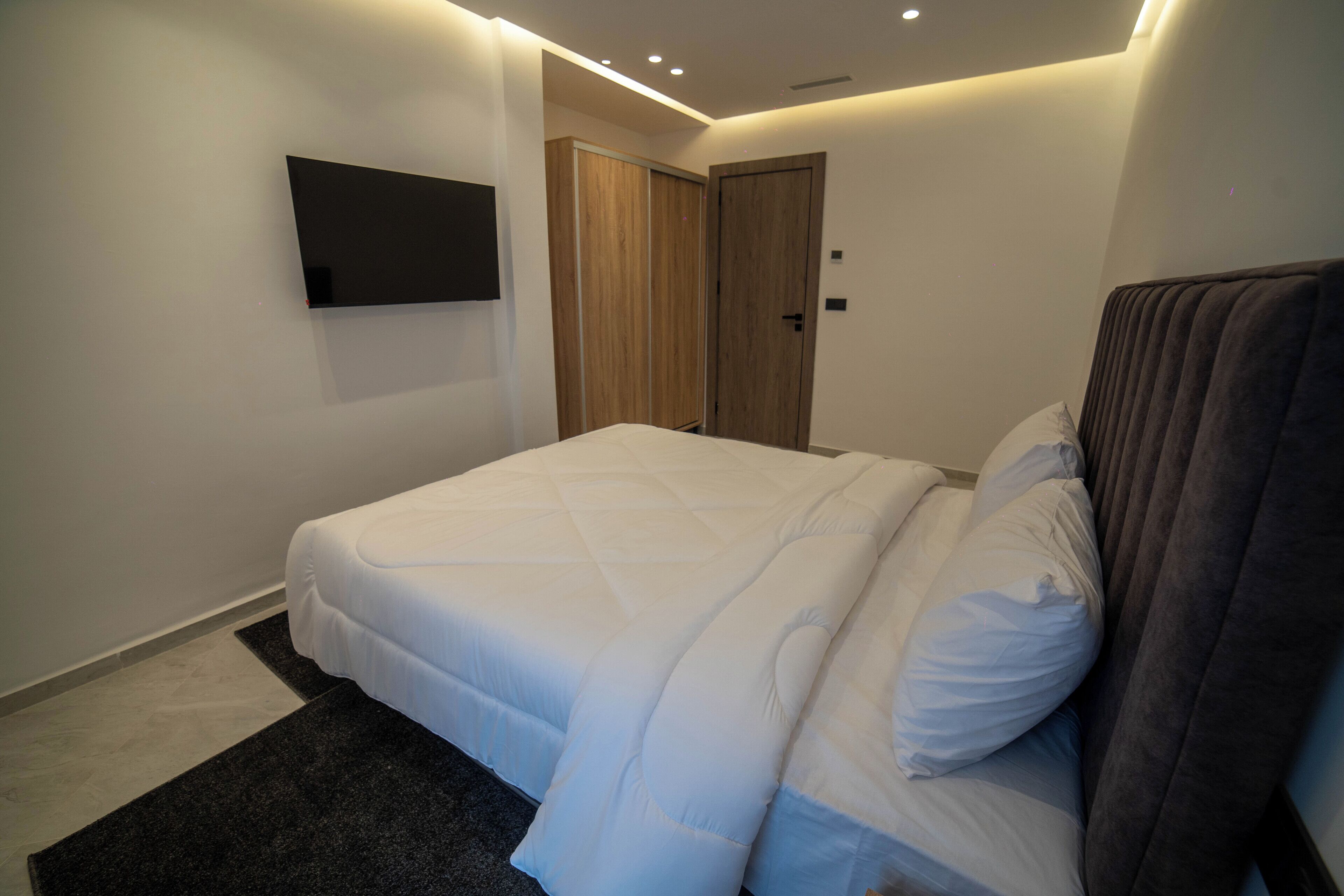 A modern bedroom with a bed, a flat-screen TV mounted on the wall, and a wooden door.