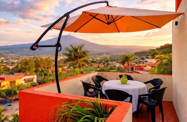 A terrace with a table and chairs, an umbrella, and a view of a town and mountains.