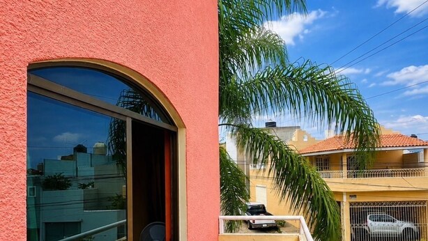 Uma varanda com chão de azulejos azuis, uma porta de vidro que dá para um quarto e vista para edifícios vizinhos e uma palmeira.