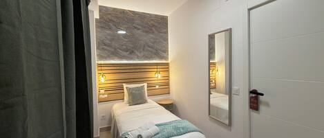 A modern hotel room with a bed, bedside table, wall-mounted lights, a mirror, and an air conditioning unit.