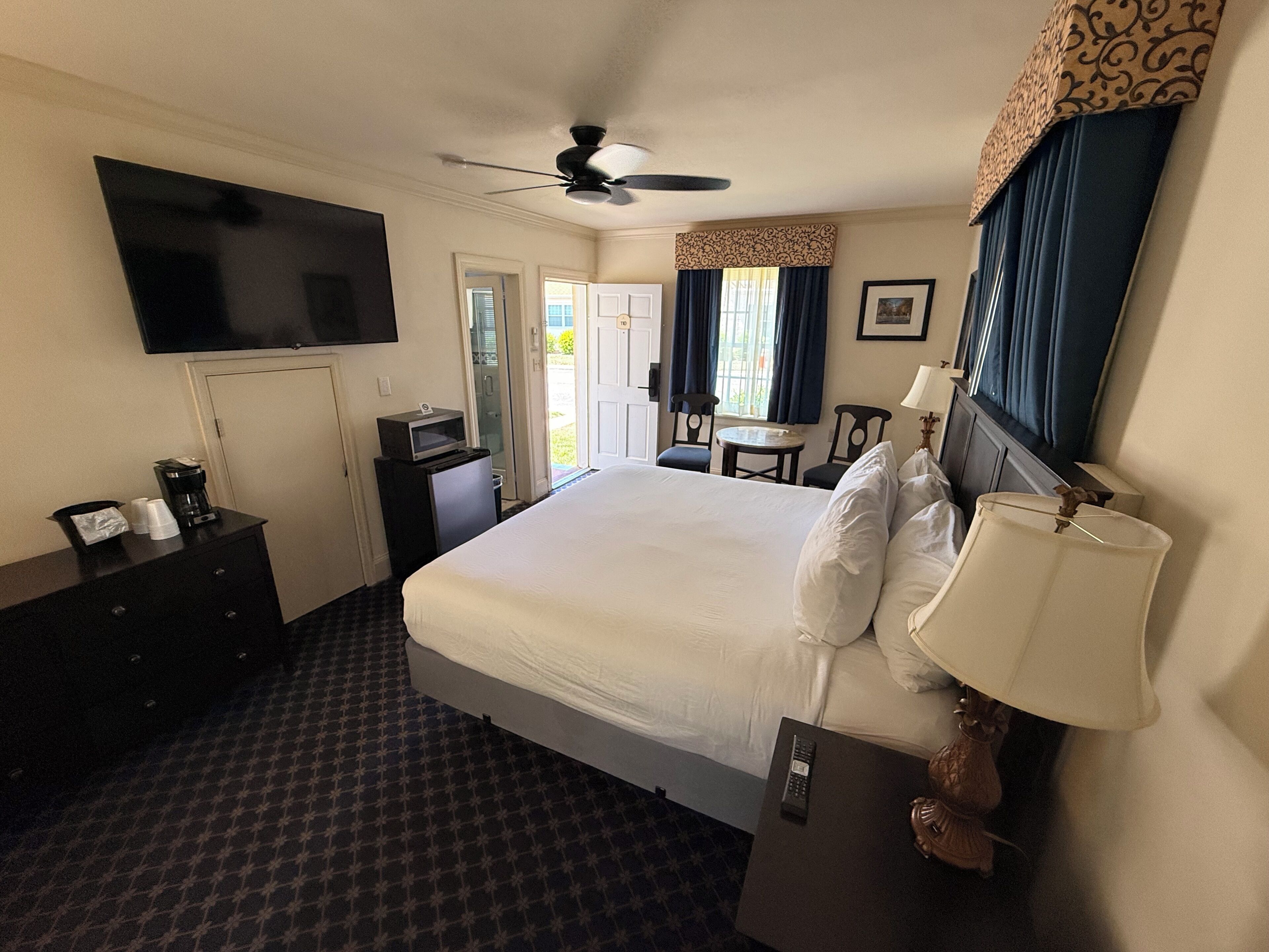 Habitación de hotel con cama, televisión, escritorio, silla, ventilador de techo y ventana con cortinas.