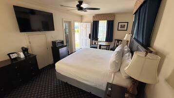Habitación de hotel con cama, televisión, escritorio, silla, ventilador de techo y ventana con cortinas.