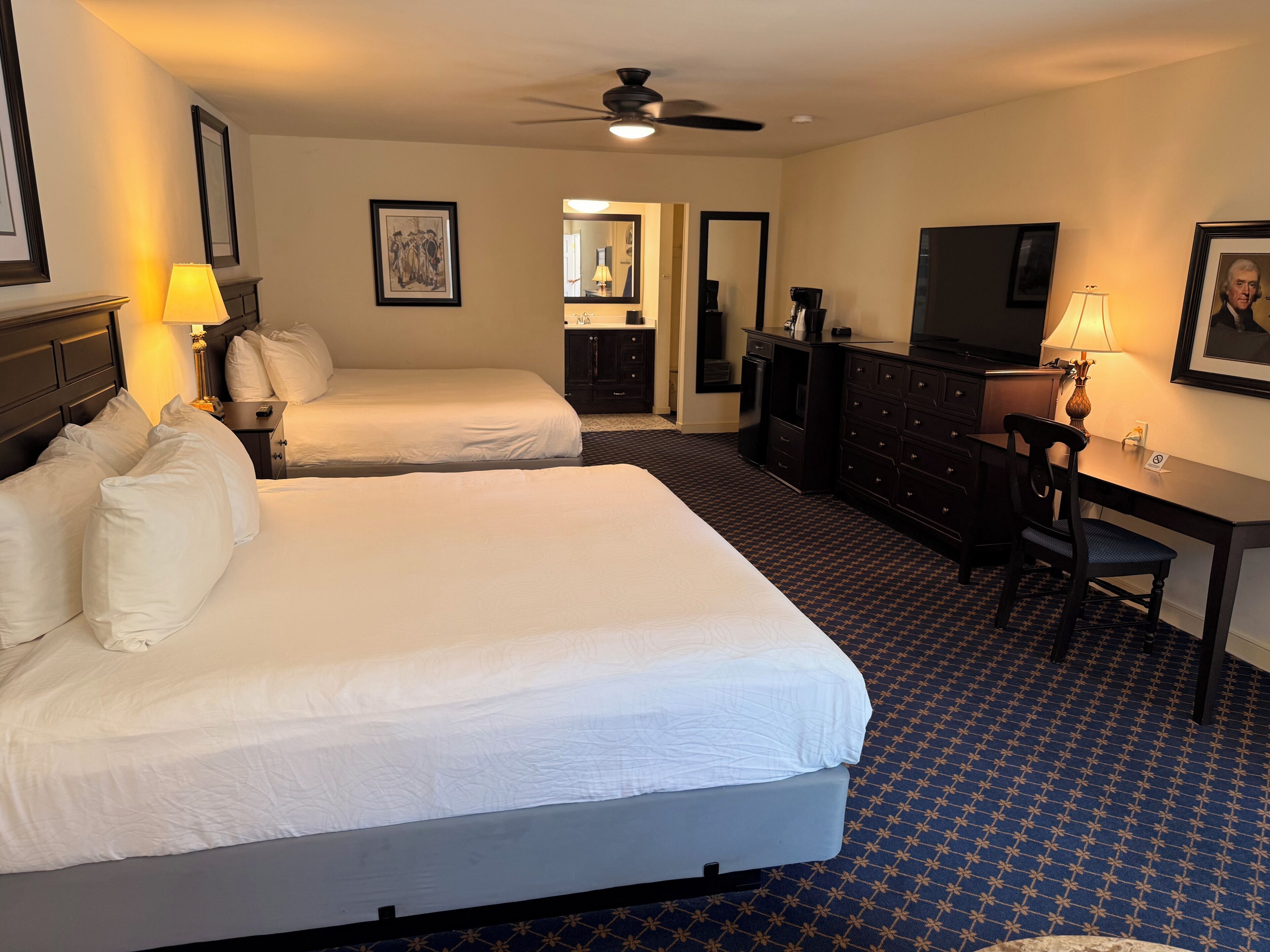 A hotel room with two beds, a desk, a chair, a television, a ceiling fan, and a bathroom visible through an open door.
