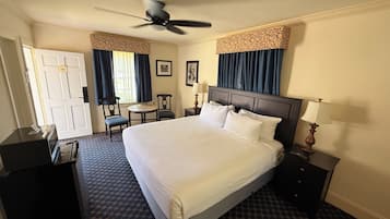 A hotel room with a large bed, a desk, a chair, a small table, a ceiling fan, and a nightstand with a lamp.