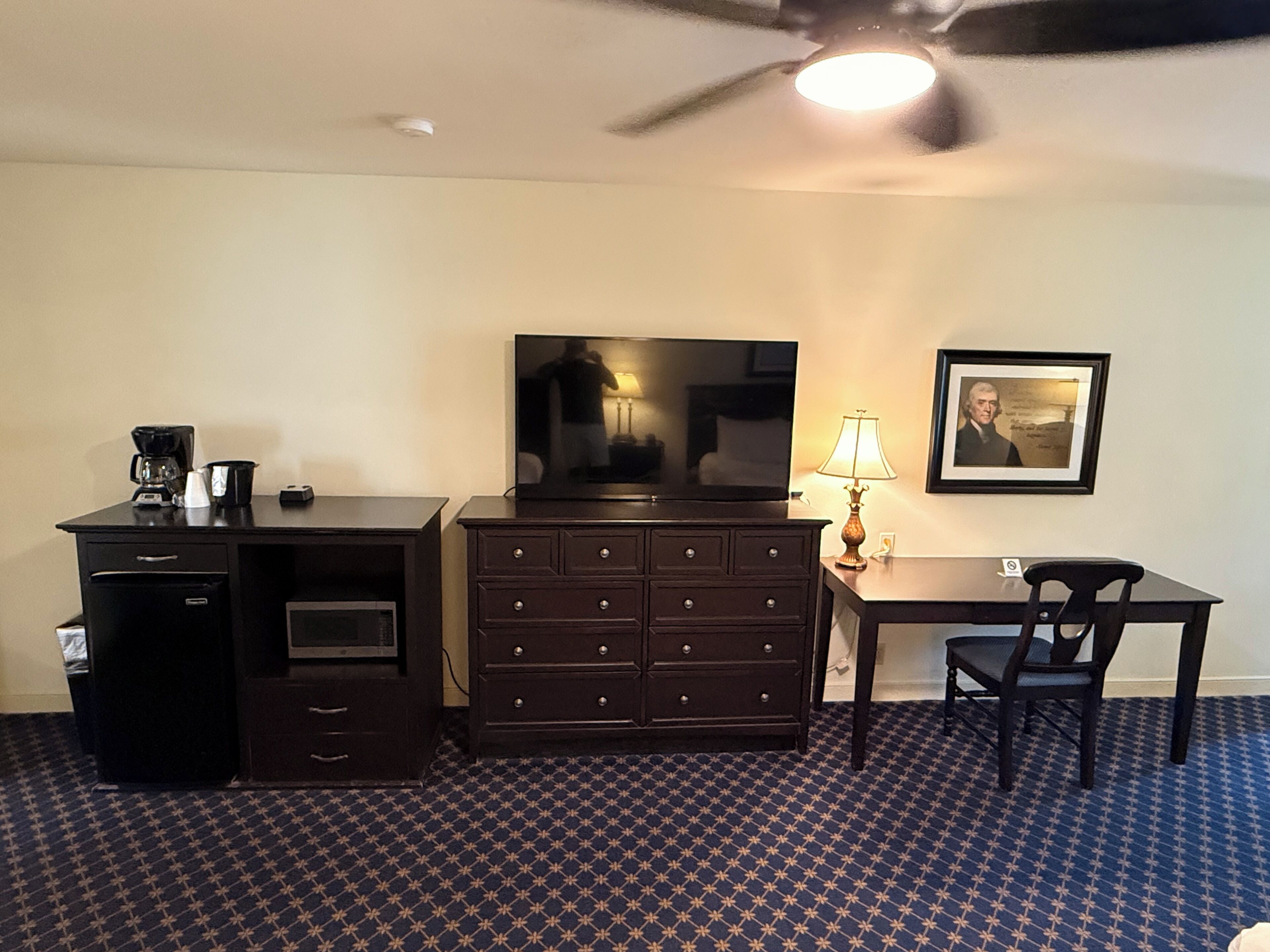 A hotel room with a desk, dresser, television, lamp, chair, and ceiling fan.