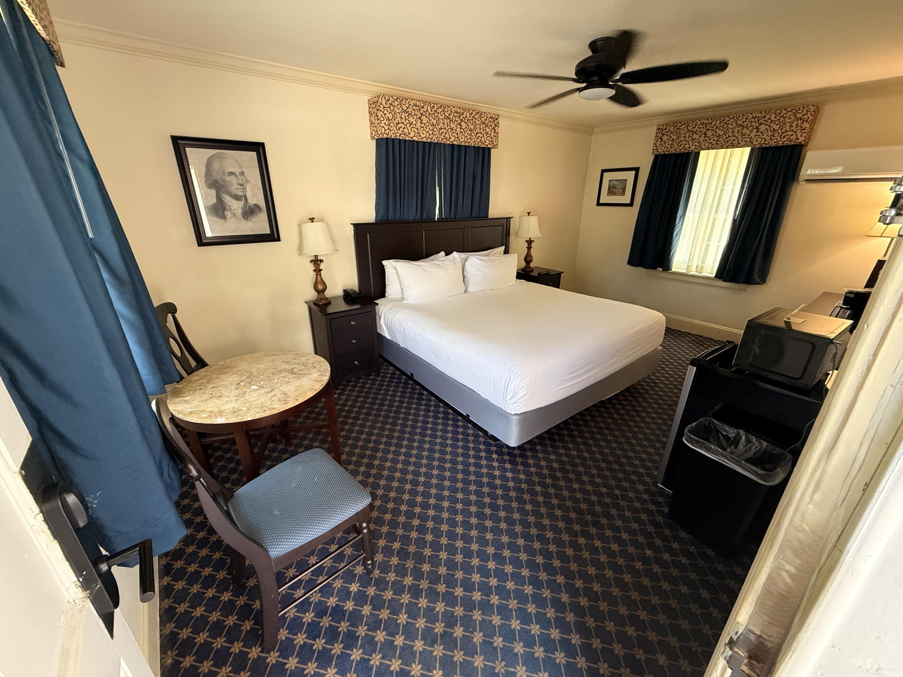 A hotel room with a bed, a desk, a chair, a ceiling fan, and a waste bin.