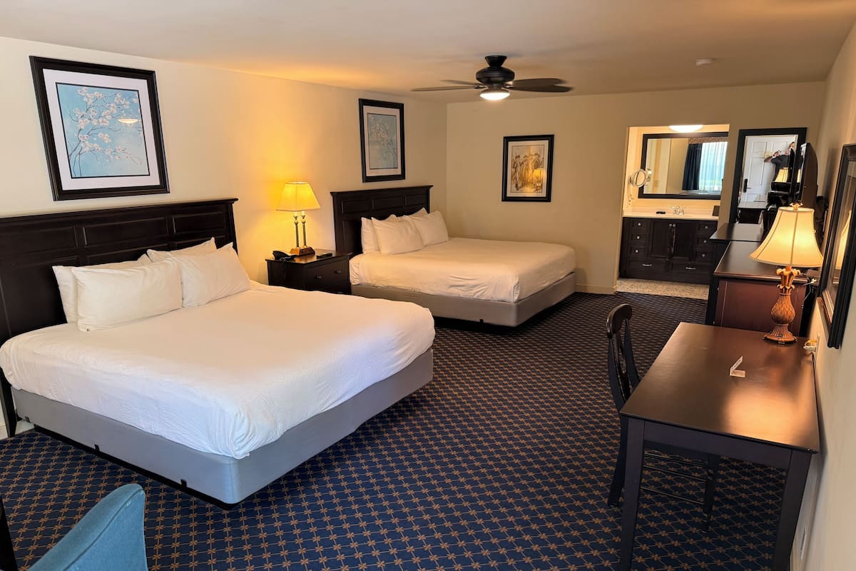 Habitación de hotel con dos camas, un escritorio, una silla, una lámpara, un ventilador de techo y cuadros en las paredes.