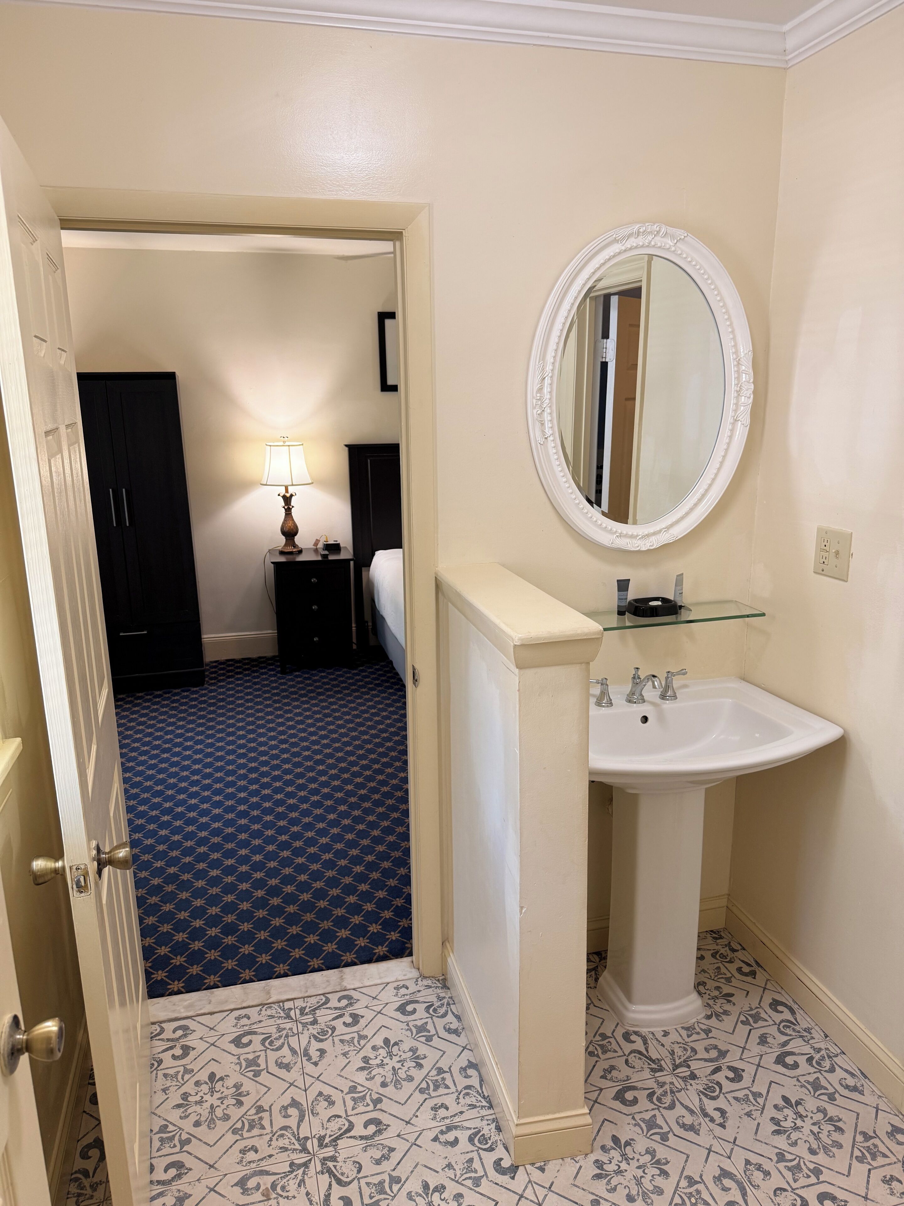 Ein Hotelbad mit einem weißen Waschbecken, einem runden Spiegel, einem blau gemusterten Boden und einer Tür, die zum Schlafzimmer führt.