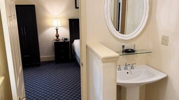 Un baño de hotel con un lavamanos blanco, un espejo redondo, un suelo azul con estampado y una puerta que da a un dormitorio.