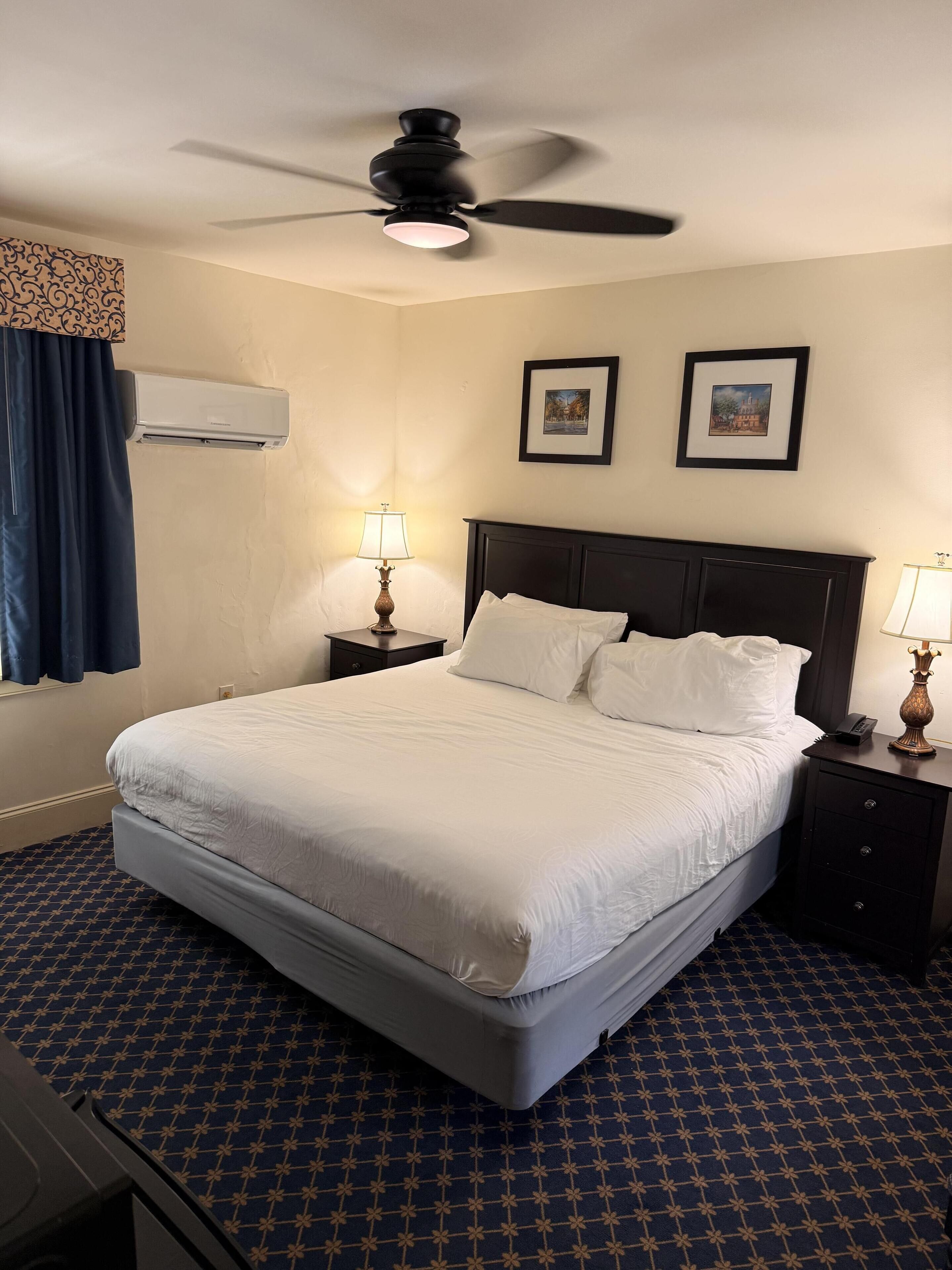 Una habitación de hotel con cama, dos mesitas de noche con lámparas, un ventilador de techo y un aire acondicionado.