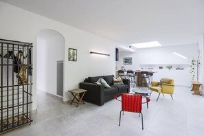A modern living room with a sofa, a red chair, a dining area, and a kitchen in the background. - Charming house between Hossegor golf and Capbreton with patio under the pines (CAPBRETON)